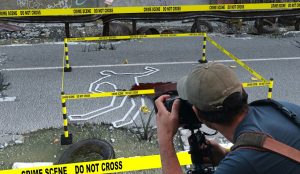 Photo illustration of a photographer documenting a homicide scene with crime scene tape surrounding a body outline marked in white tape on the ground next to a pool of blood staining the road.