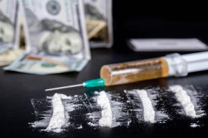 Photograph depicting powdered drugs and drug paraphernalia, with a pile of money blurred in the background.