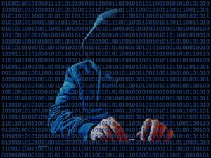 An image of a faceless hacker in a dark hooded sweatshirt sitting and typing at a keyboard, with binary code running across the foreground.