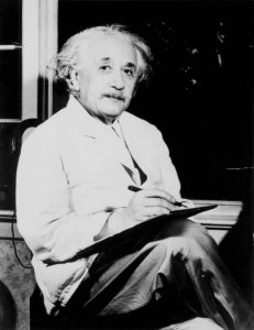 Albert Einstein dressed in white labcoat sitting in chair holding clipboard.