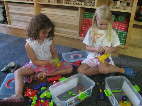 Two girls sit together on the floor of a block area in a classroom surrounded by small plastic toys.