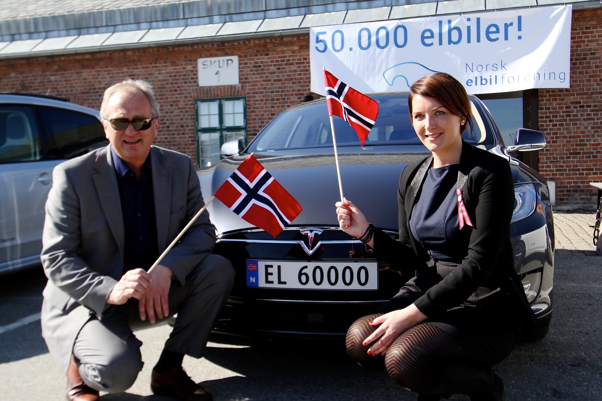 An older man standing next to a younger woman. They are both holding Norwegian flags next to a Tesla car.