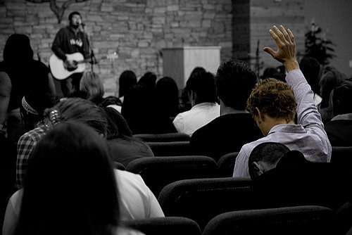 Members of a church listening to a man play guitar and sing. A singular man raises his hand in praise