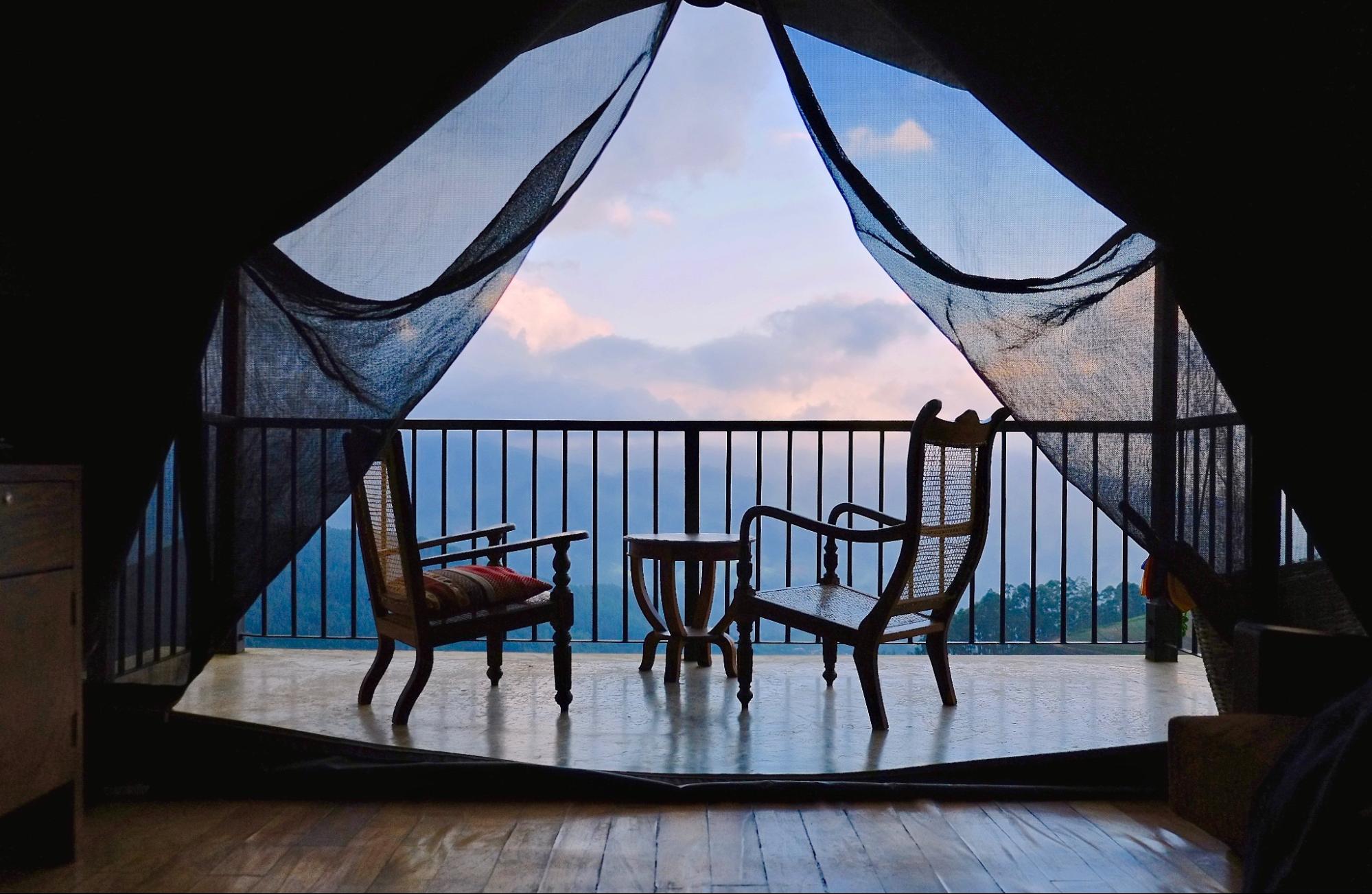 A balcony with custom wooden furniture overlooks clouds and trees. This is a view from the inside of a glamping site.
