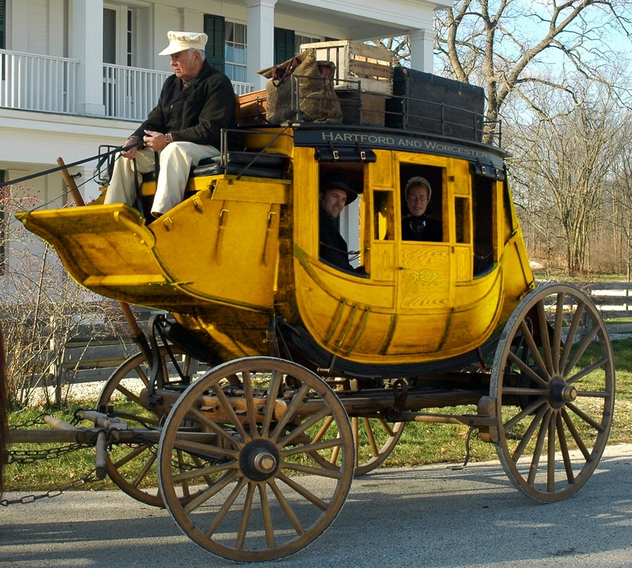 An old Sturbridge village with a yellow stagecoach, a man and woman riding inside.