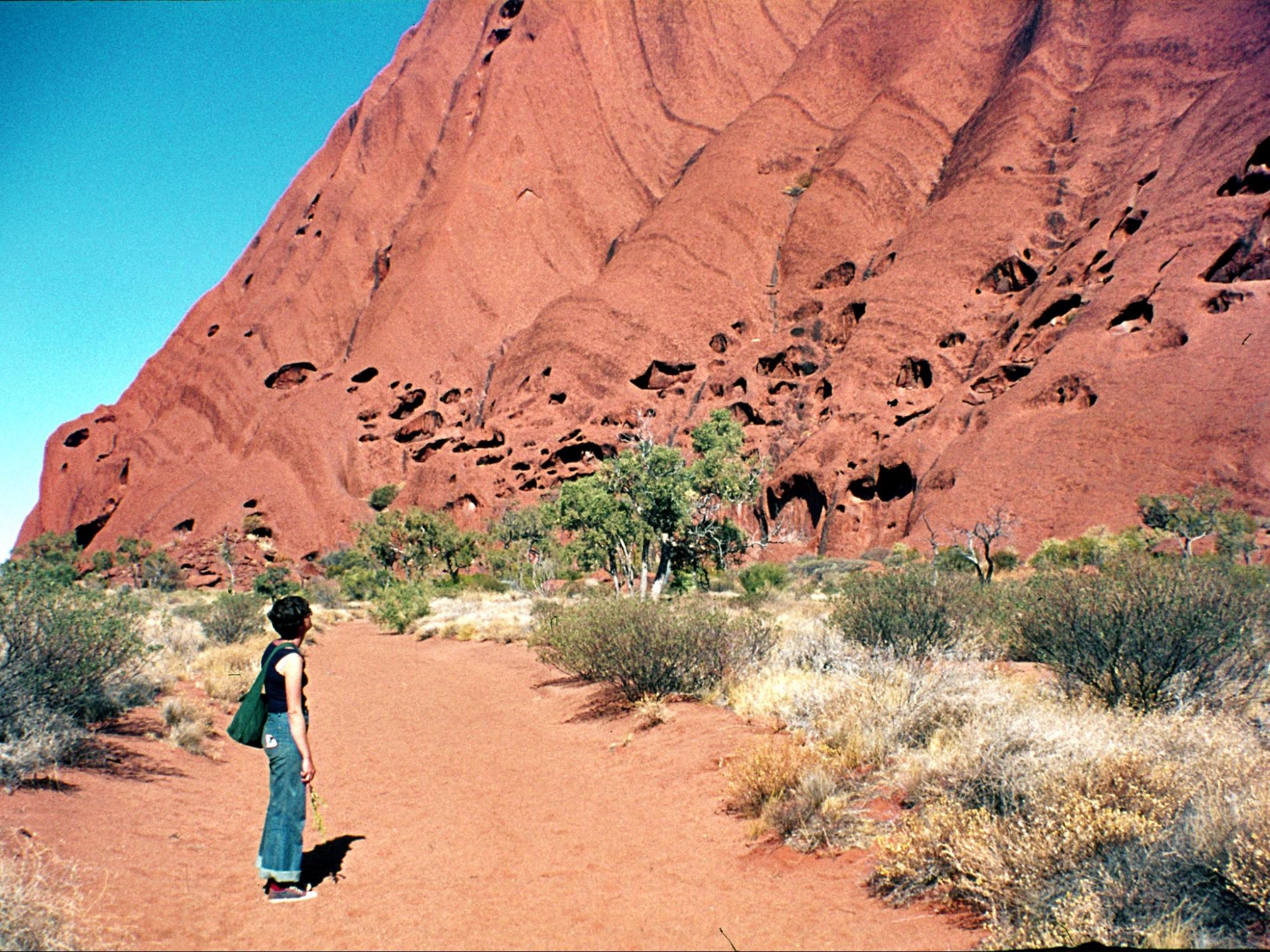a woman with short dark hair, a black short sleeved shirt, and jeans, stands at the base of Uluru gazing up its rockface. The landscape is scrubland and a red sandy pathway where the women stands. Uluru is a large, red colored rock.