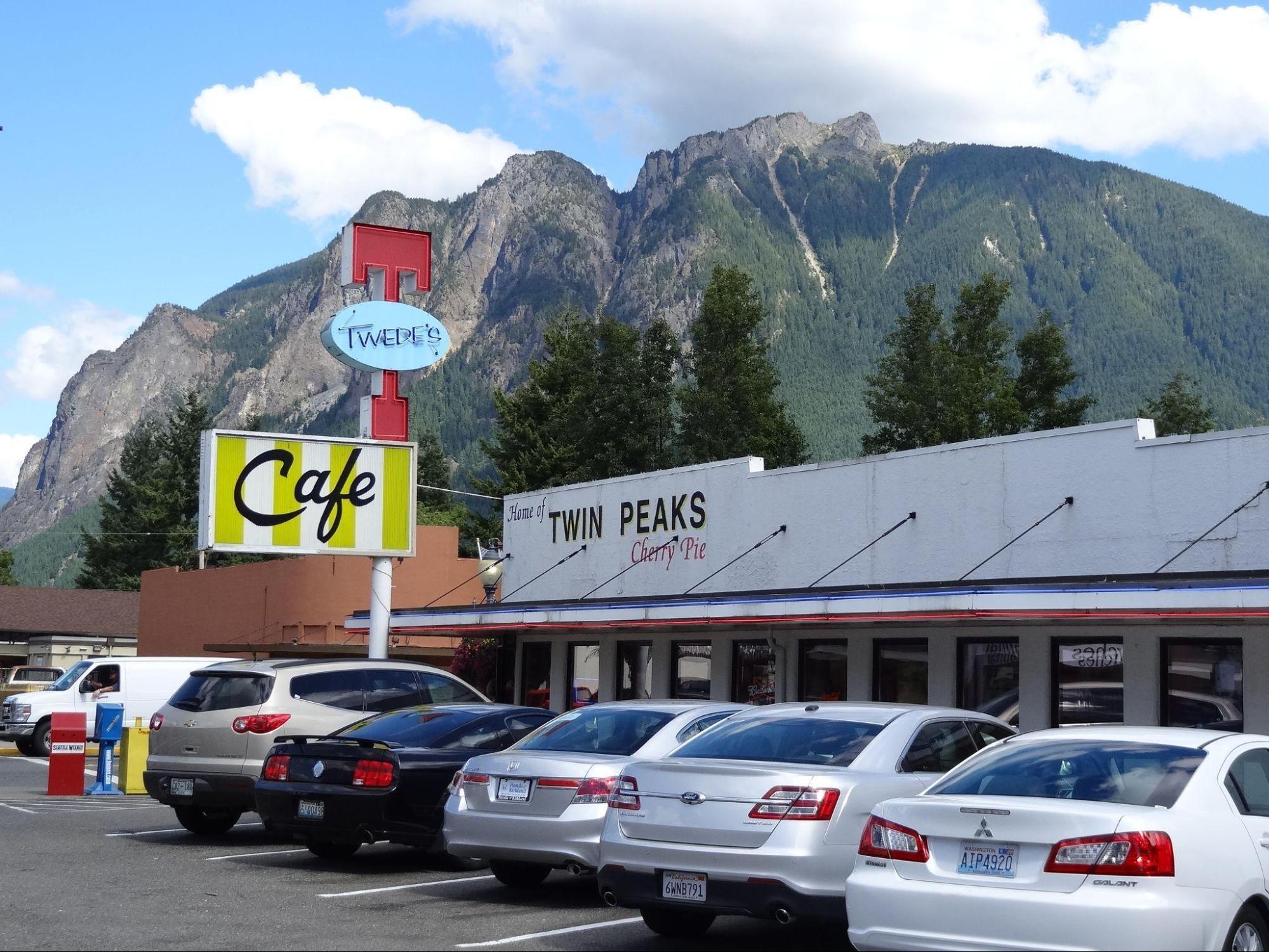 Photograph depicts the outside of a cafe with the words, “Twin Peaks Cherry Pie” painted on the side. A sign reads, “Twede’s Cafe”. Many cars are parked in front of the cafe.