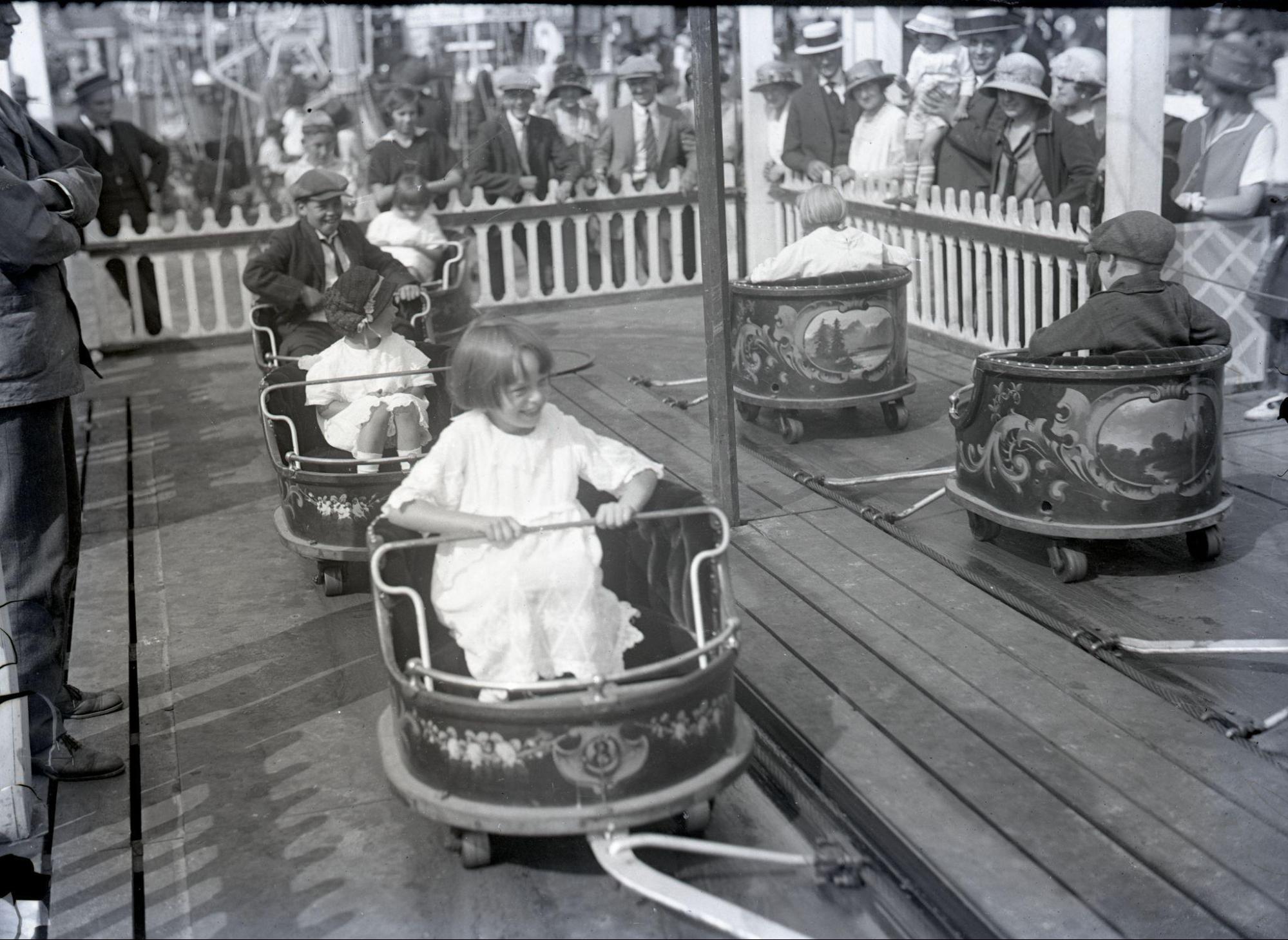 A photograph depicting a ride at Coney Island. A girl in a white dress sits in a cart. Multiple other children sit in their own carts. The carts are attached to a track. The photograph is in black and white.