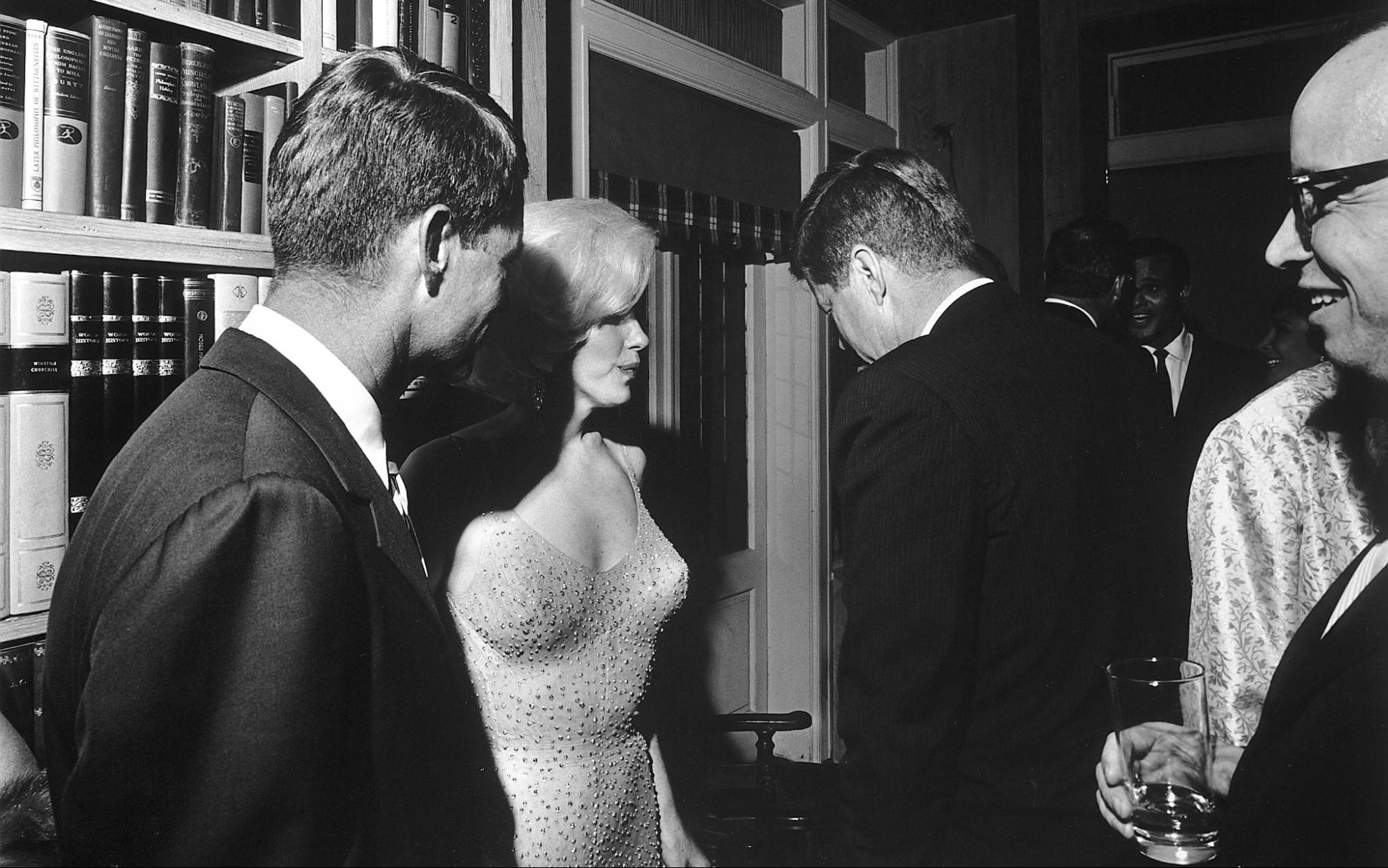 Many men stand in a room around Marilyn Monroe, who is wearing a low cut, light colored, fitted dress. Photograph is in black and white.