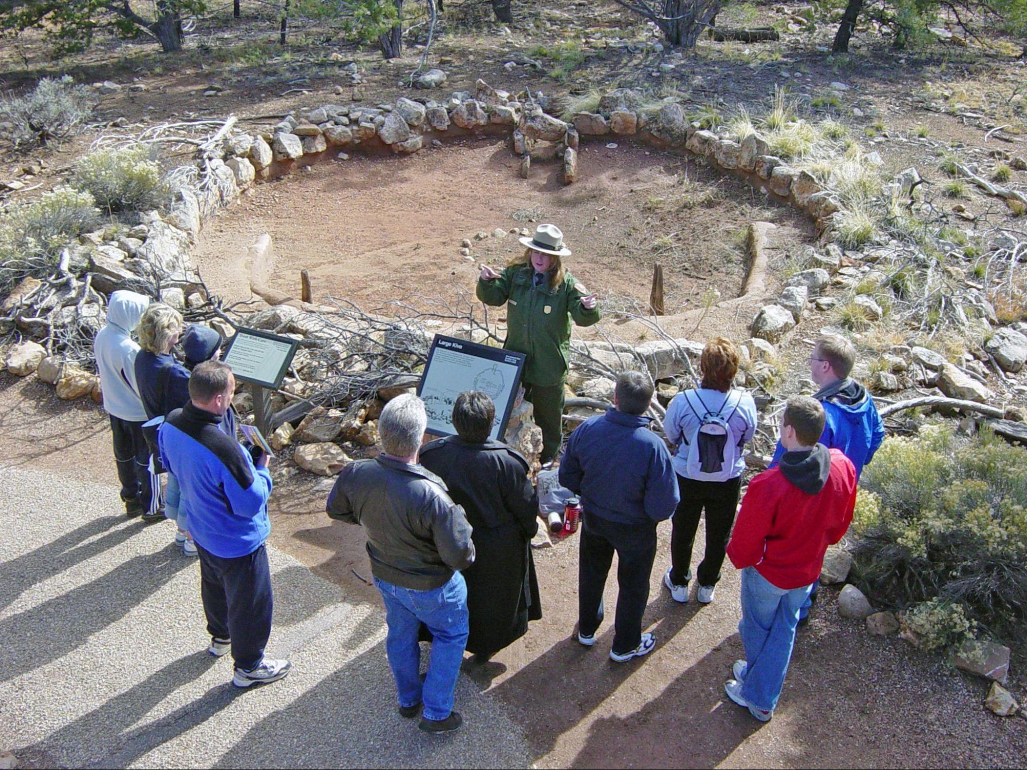 A female national park ranger speaks to a group of people who stand around an ancient ruin–a circle of earth surrounded by rocks.