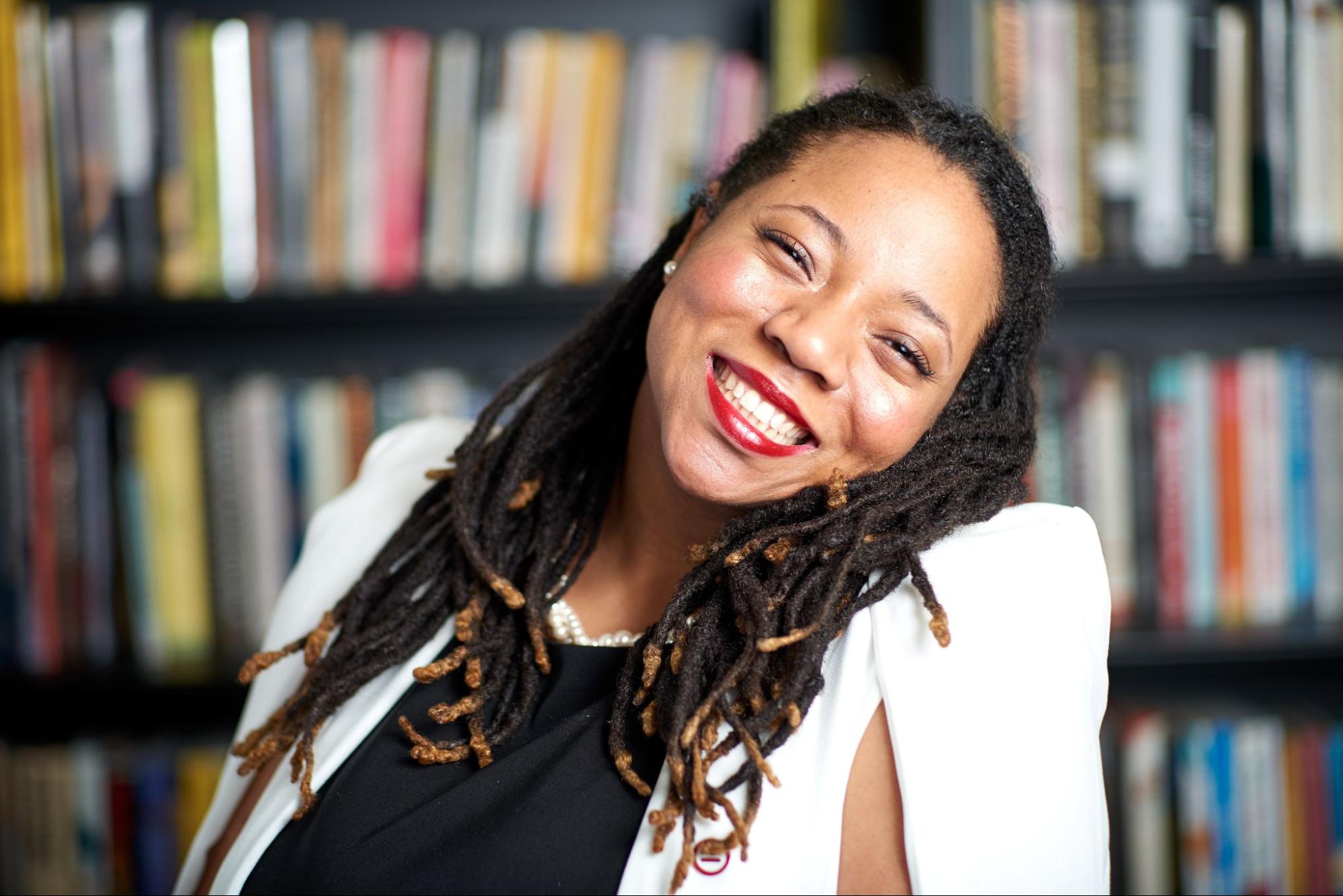 A photograph depicting a woman with her head tilted to the right. She smiles wide. She has black dreadlocks with lighter colored tips.