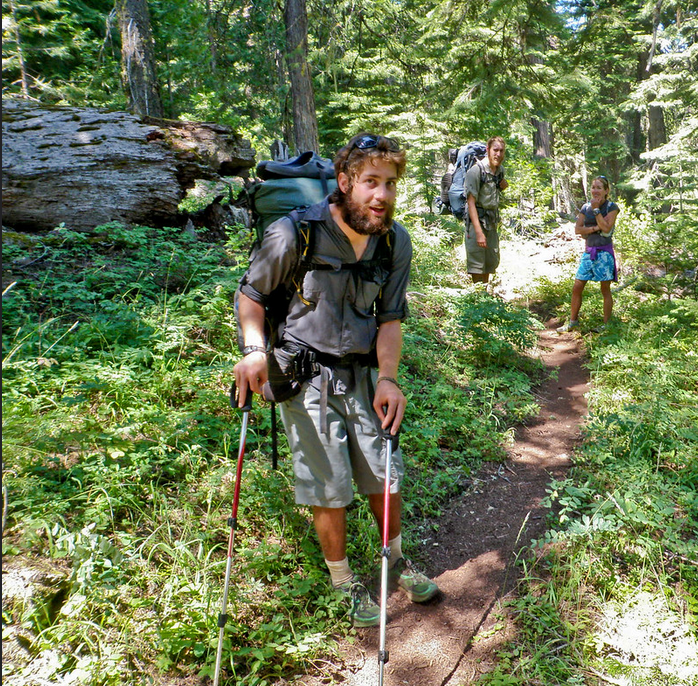 Three hikers face the camera on the Pacific Crest Trail. Two of the hikers carry hiking packs, and one of them is using walking sticks.