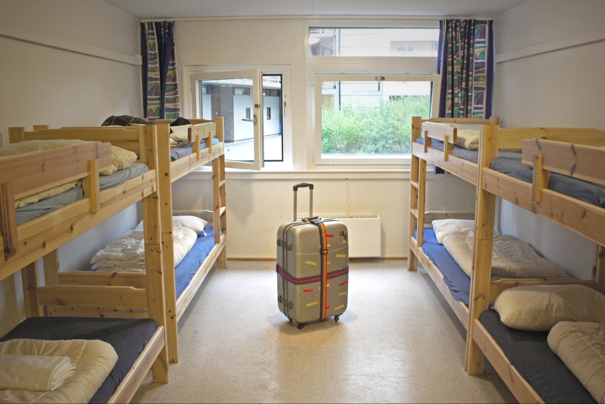 A hostel shows four bunk beds with folded sheets and pillows. A carry-on sized luggage stands in the middle of the room.