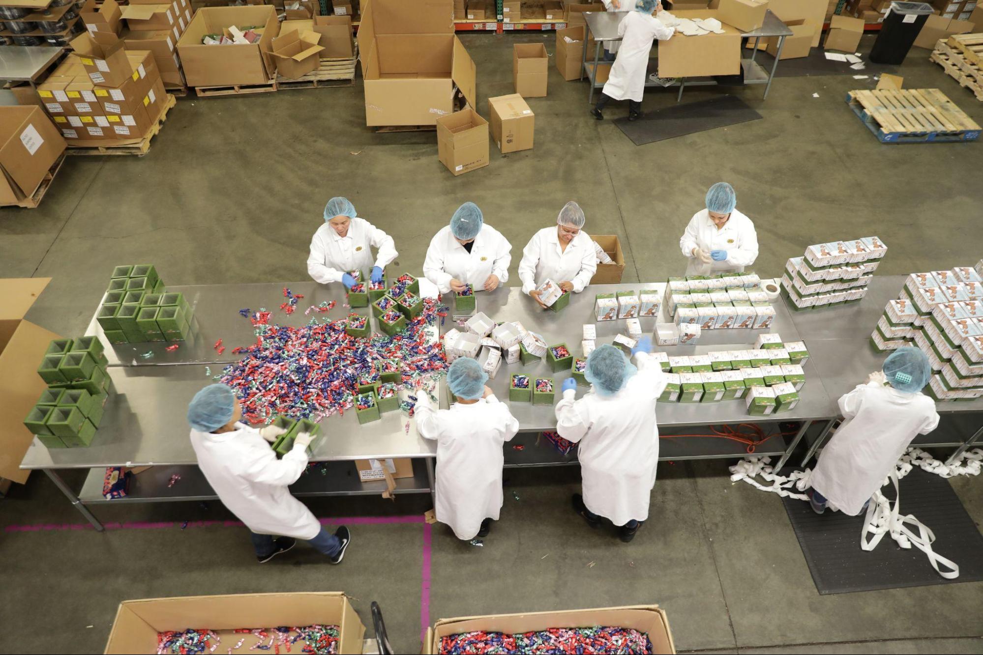 Eight workers in white lab coats and hairnets stand around a steel table sorting colorful truffles into boxes.