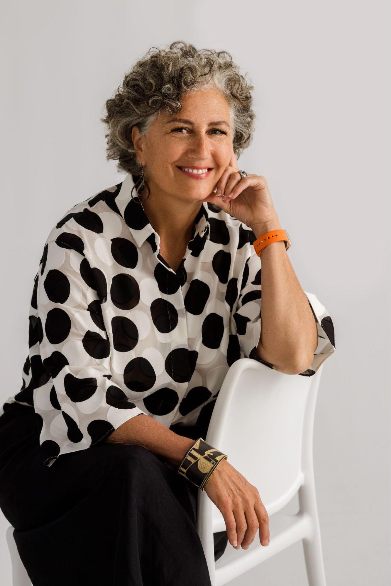A woman with short, curly hair in a white shirt with black dots and black pants sits on a white chair smiling