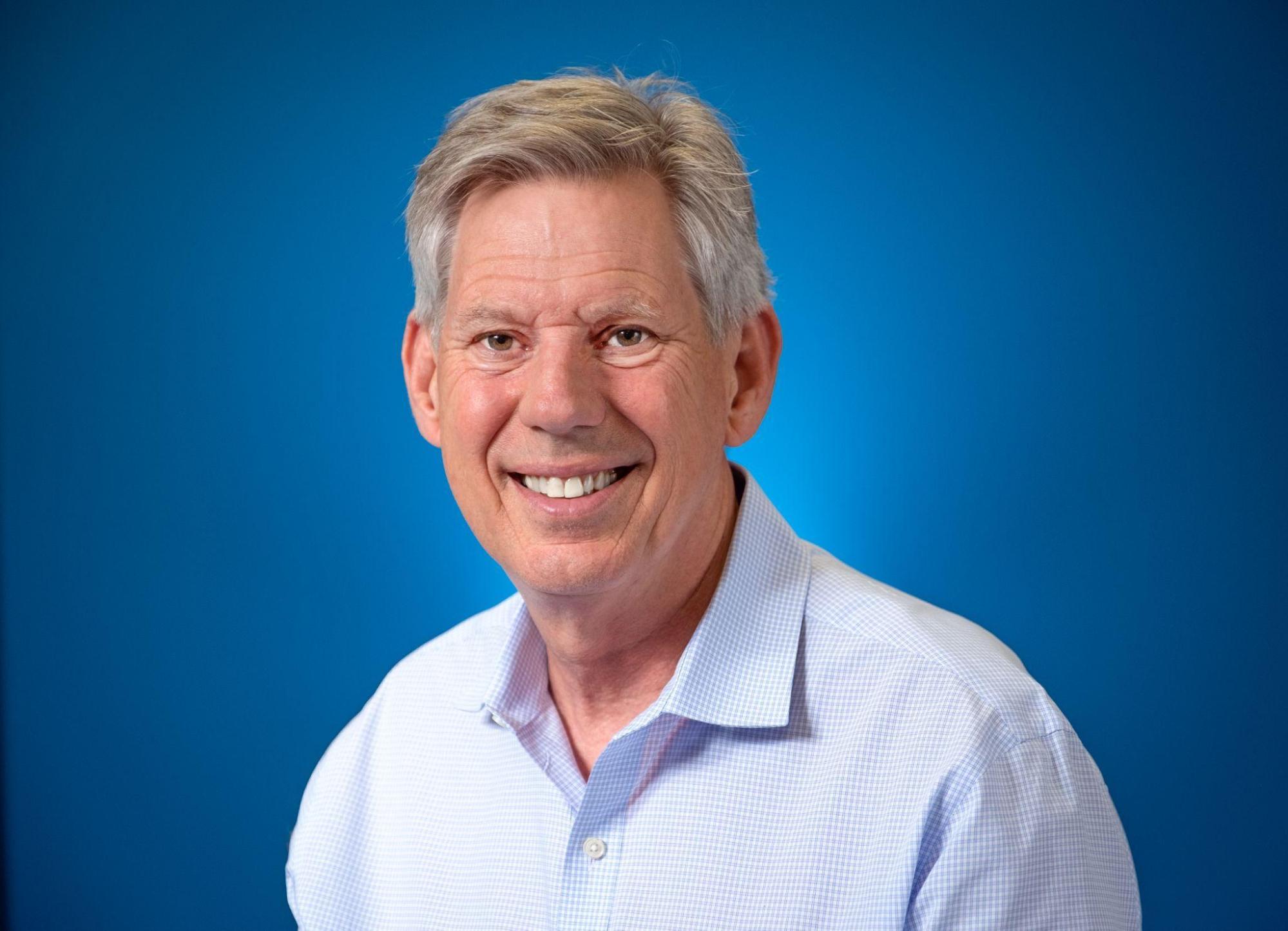 A male figure with gray hair and a light colored collared shirt is facing the camera and smiling.