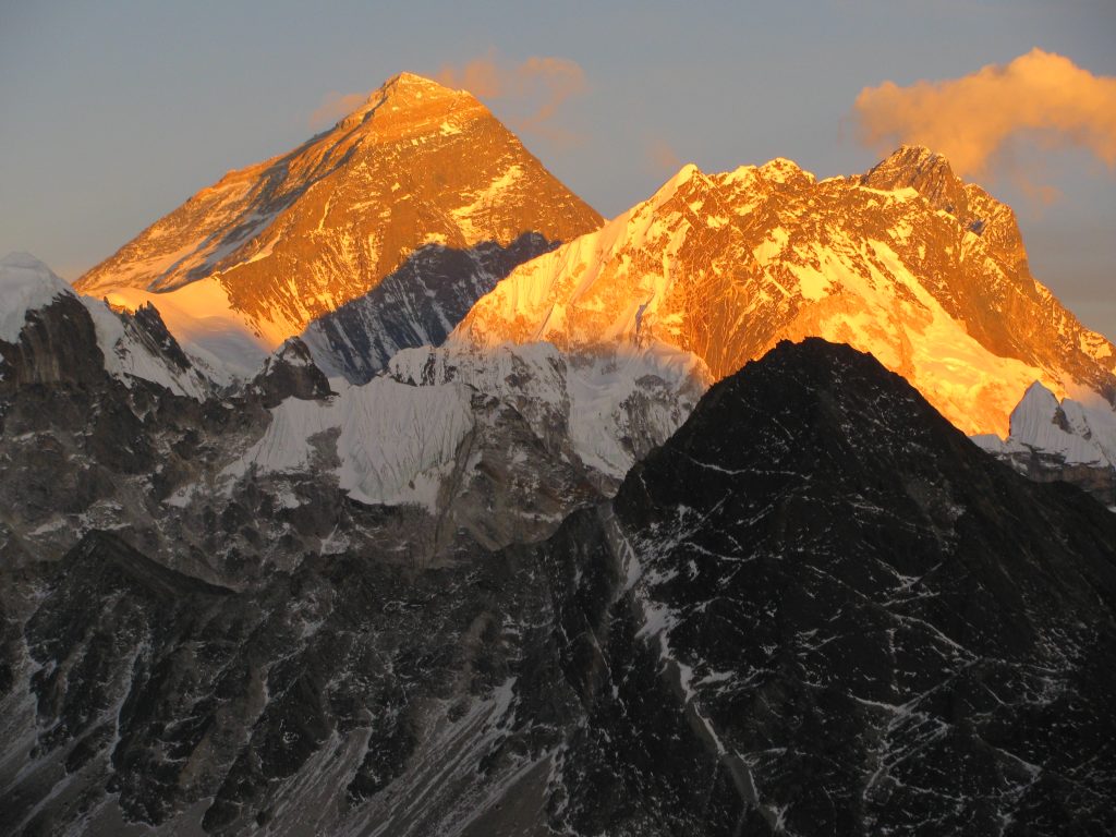 A photo of the peak of Mount Everest. Most of the mountain is in shadow, but the peak itself is bathed in yellow-orange sunlight.