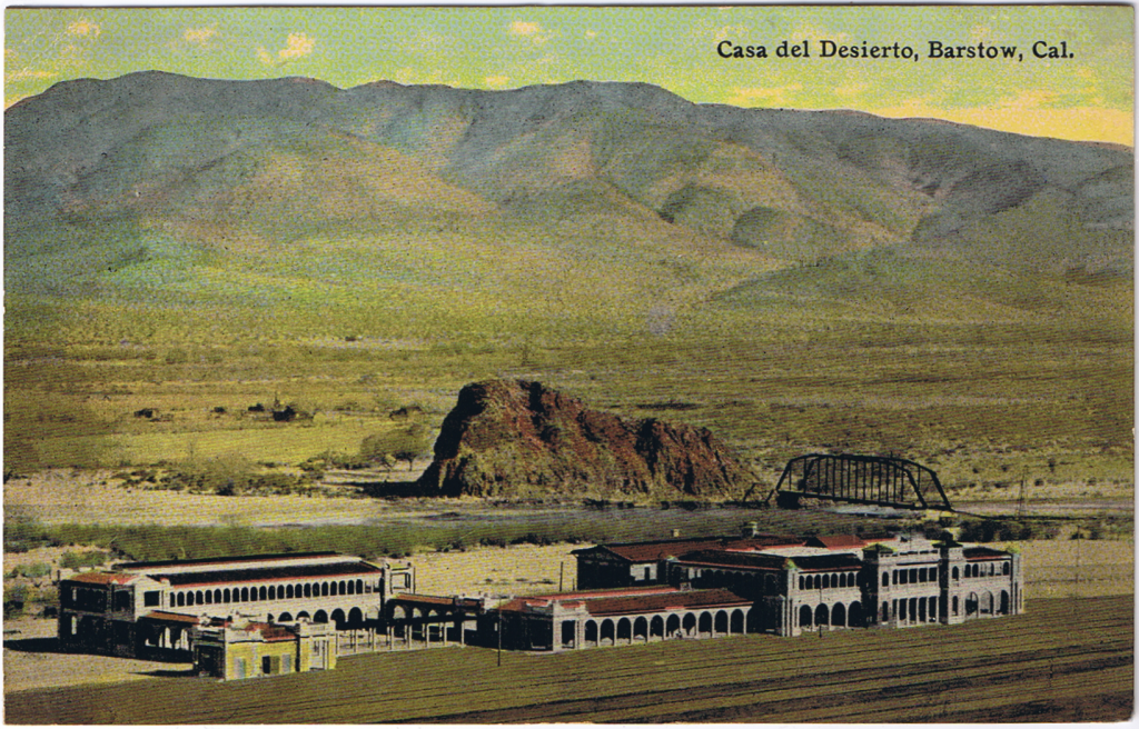 A grainy but colorized photo postcard, featuring a white building with constant archways between a railroad and a river. It looks small in comparison to the desolate landscape behind it, which takes up most of the photo.