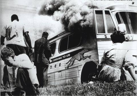 A black and white photograph of a Greyhound bus, its door open and smoke billowing out. Four Black passengers are visible, two sitting and two standing, in nice outfits, looking unharmed but tired.