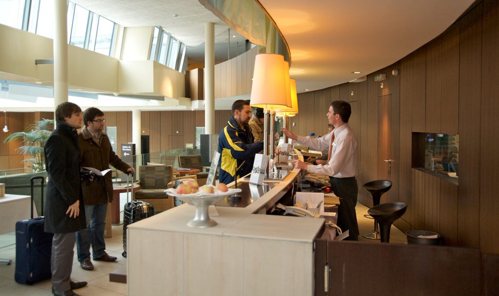 Several travelers with luggage wait their turn at a long, curving hotel front desk.