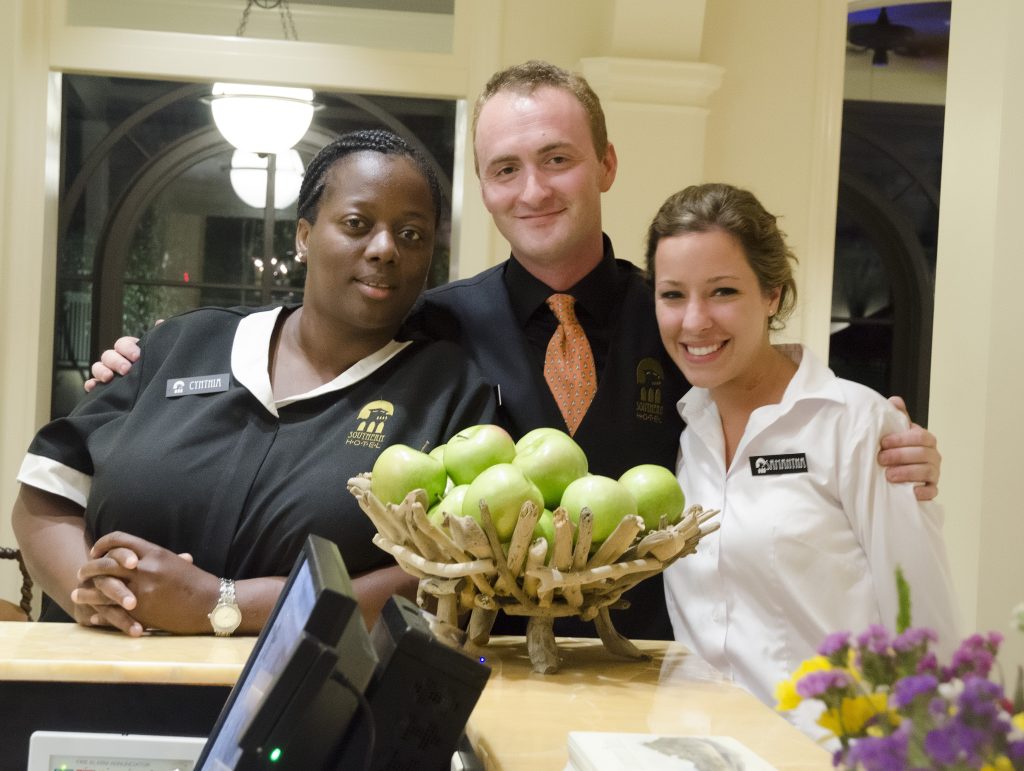 Three people in different hotel uniforms pose for the camera, smiling, behind a large bowl of apples.