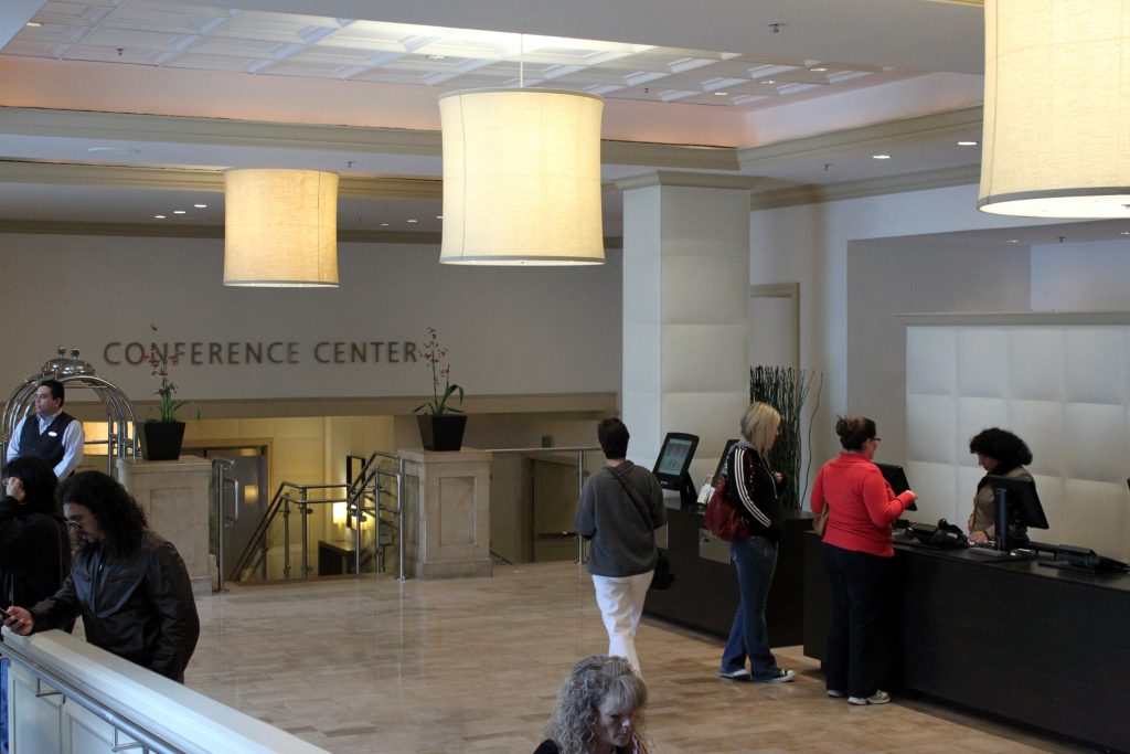 A variety of travelers move through the lobby of a hotel, while two engage with an employee at the reception desk.