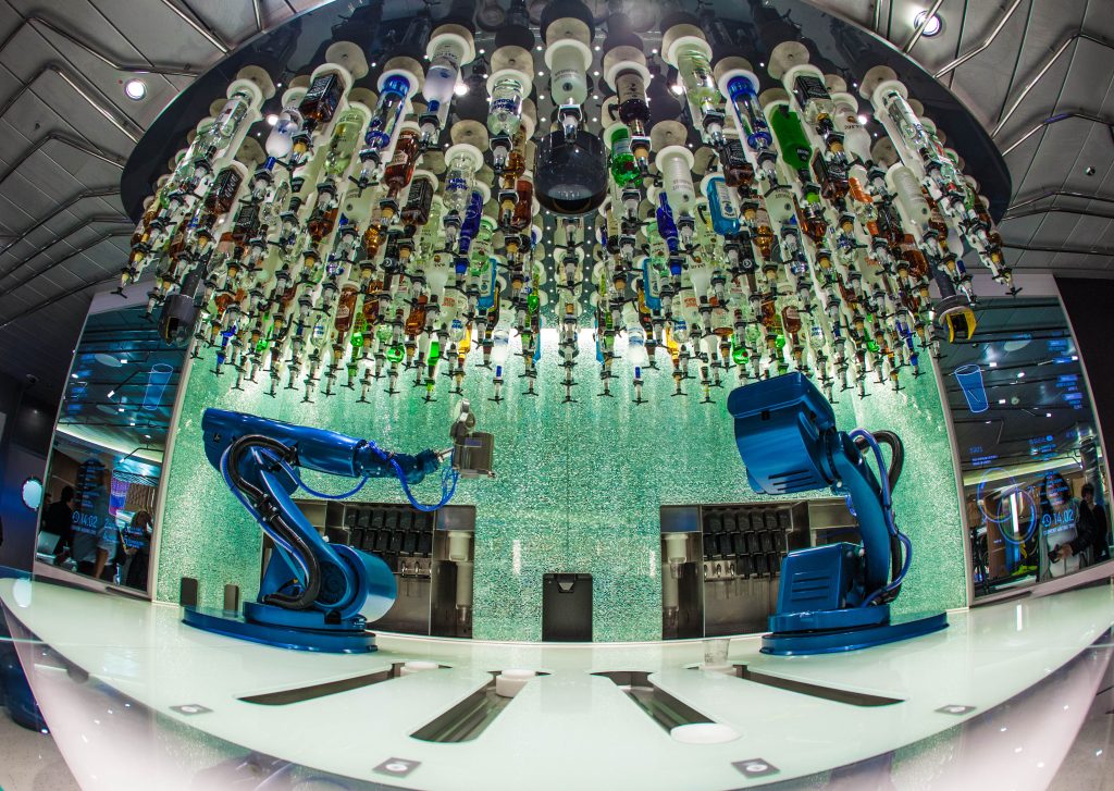 Two metallic blue robots prepare drinks on a bartop, with alcoholic bottles upside down on the ceiling above them.