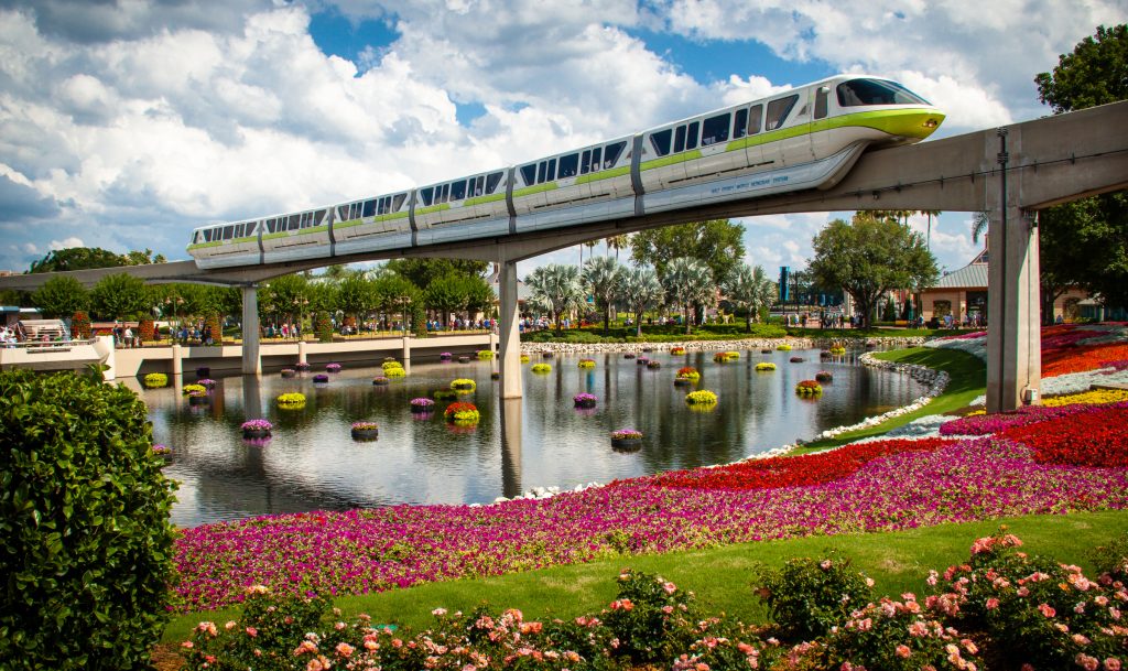 A white-and-green monorail train on a narrow, concrete bridge passes over a pond and its near bank of colorful flowers.