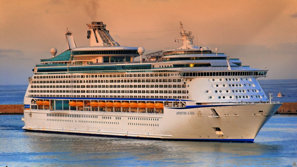 A squat, white cruise ship with orange emergency floats sails in calm waters, with the sky shifting from grey to peach behind it as the sun rises.