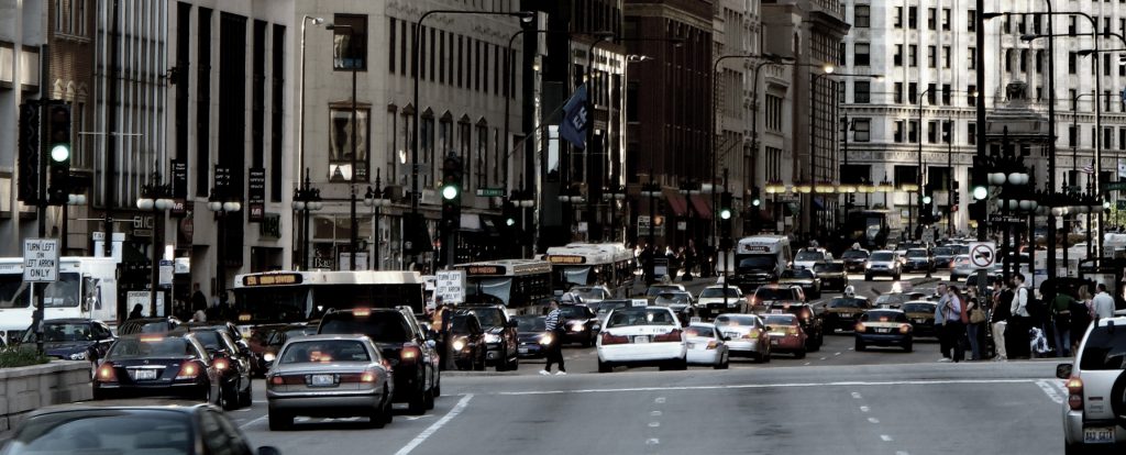A tightly-packed street features cars, buses, and taxis with their brake lights on, pedestrians moving in a crossway, and tall buildings framing the far side of the street.