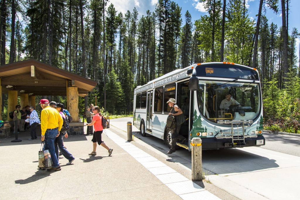 Passengers unload from a Glacier National Park shuttle bus at a wooden bus shelter, with tall trees behind.