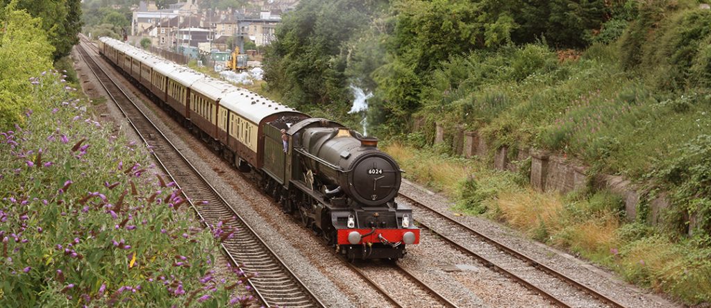 An old-fashioned train engine, attached to a coal car and multiple passenger cars, passes by a green and flowery countryside.