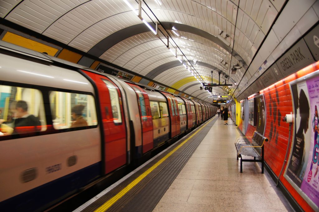 A London subway train passes by in a blur at an underground station with a white, curving roof and colorful, ad-lined walls.
