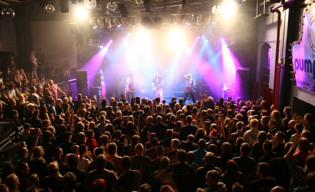 A rock band, under pink and blue stage lights, faces a packed crowd in a small, dark, indoors venue.