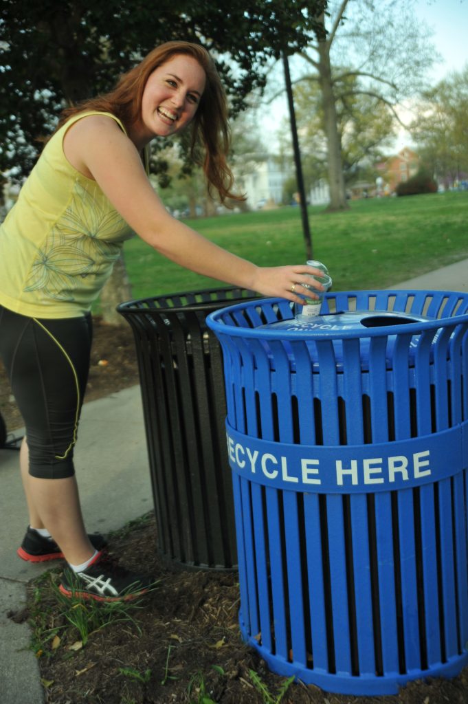 A smiling woman places a bottle in a blue recycling receptacle in a public park.