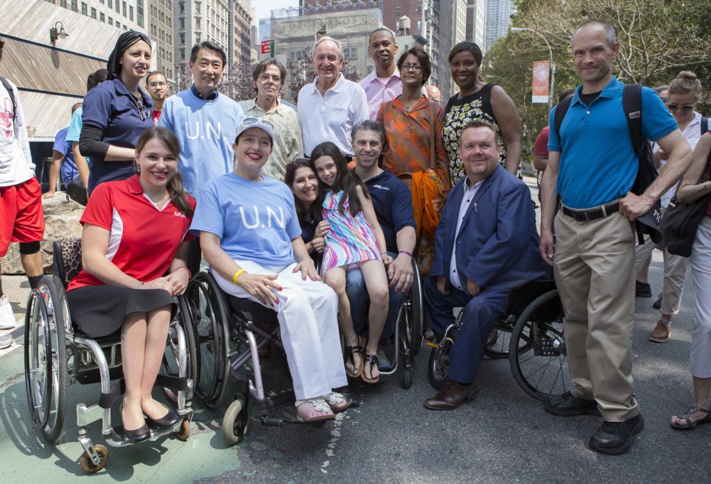 A diverse group of people, most with visible disabilities, smile at the camera on a New York City street.