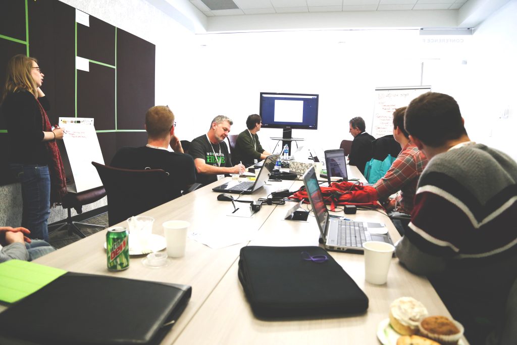 A small group of people sit around a conference table cluttered with laptops and drinks, talking and viewing a display at the far end of the table.