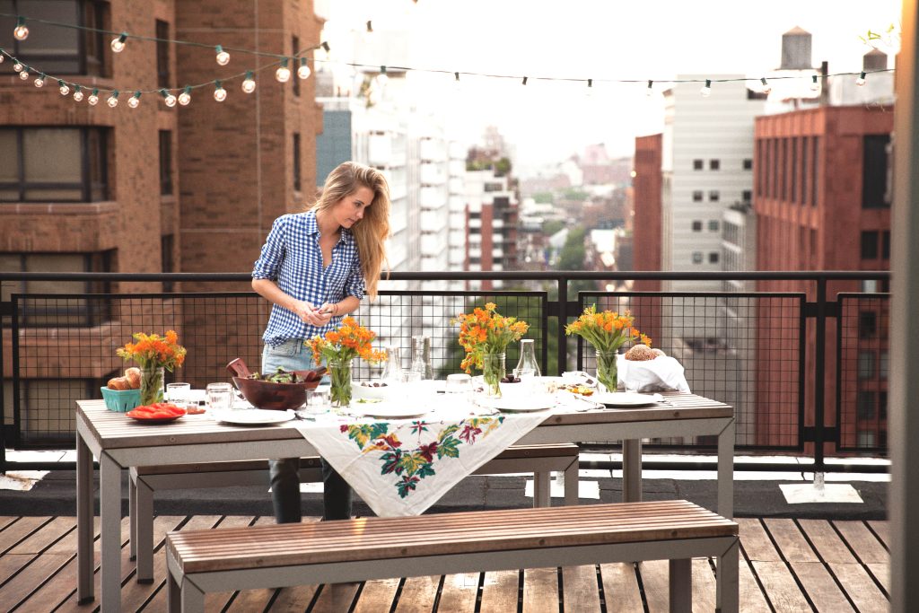 A woman looks over a table neatly set with flowers, food, and tableware, on a patio overlooking a city street.