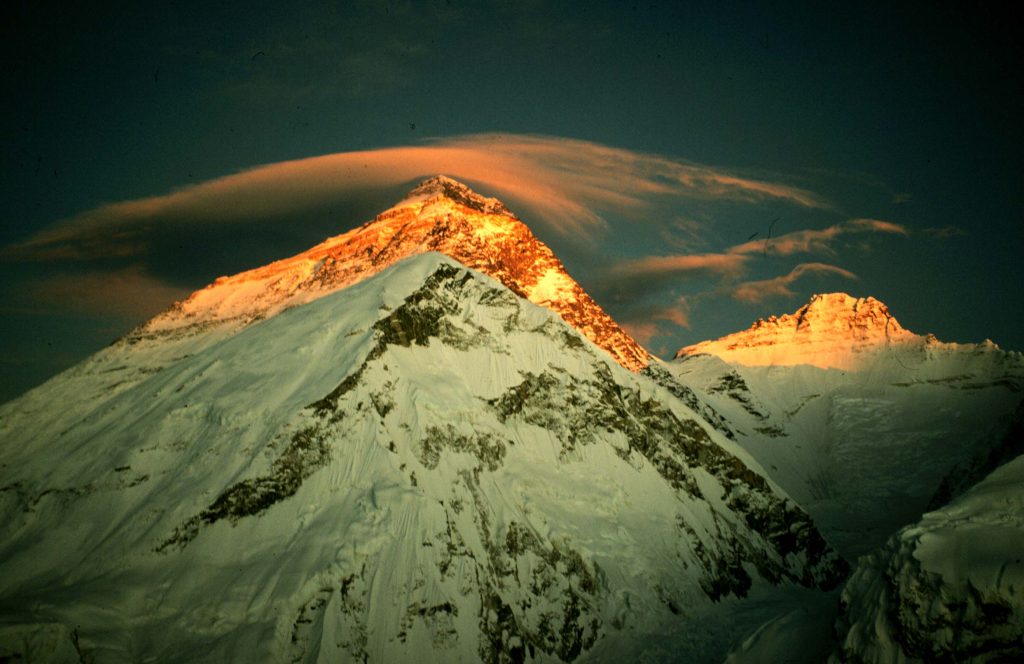 A photo of the peak of Mount Everest. Most of the mountain is in shadow, but the peak itself and several clouds drifting over it are bathed in yellow-orange sunlight.