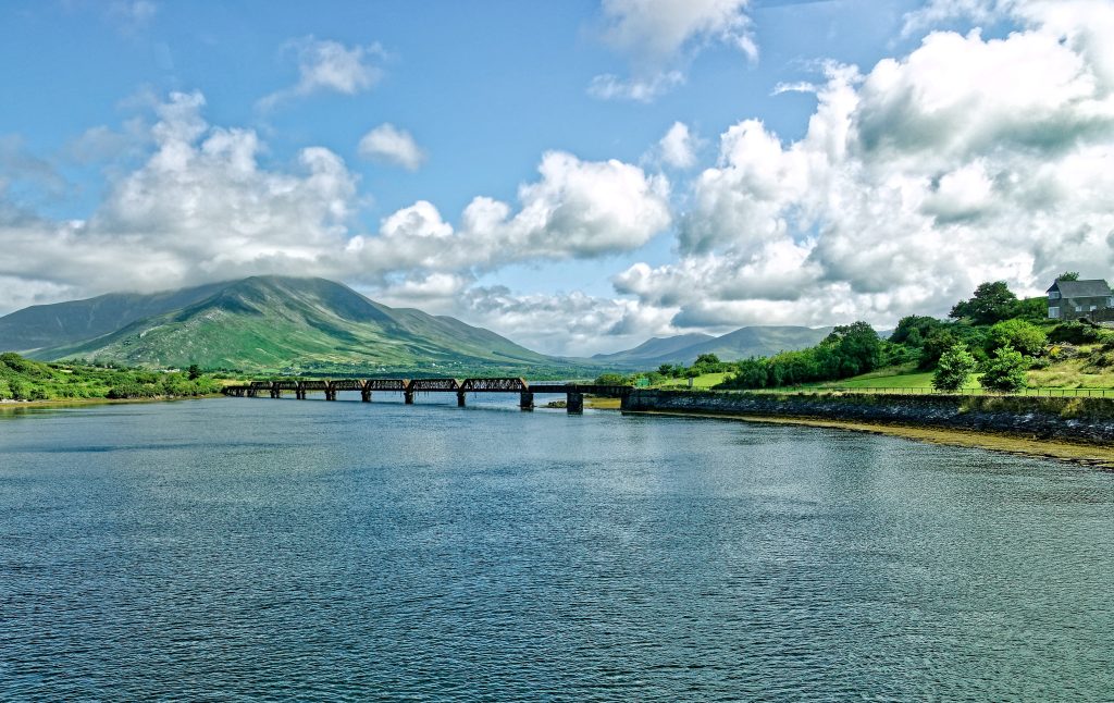 The broad expanse of a river dominates the photo, with a small, distant bridge and green mountains visible against a cloudy blue sky.