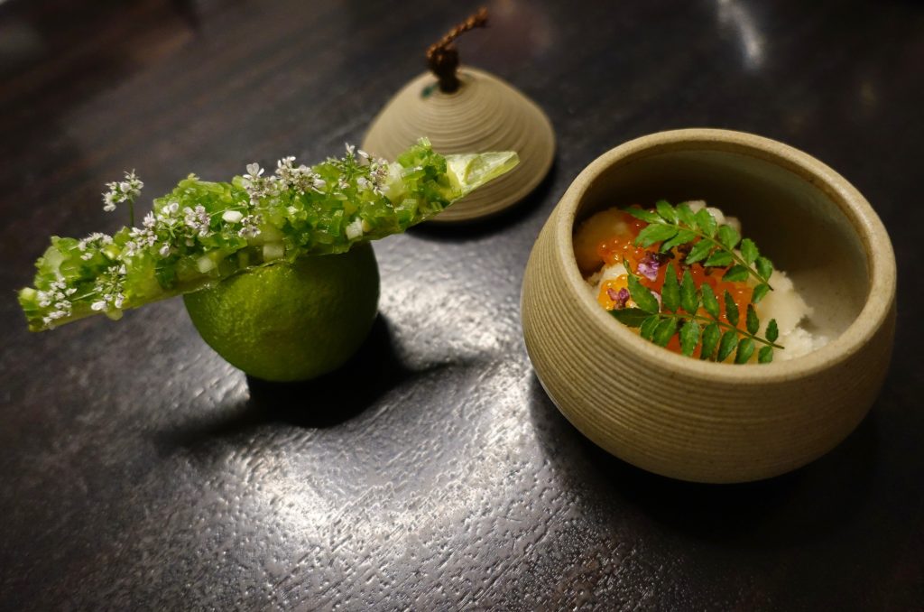 A leaf with finely chopped greens balances on top of a lime, next to a small clay pot holding a serving of caviar with delicate garnishes.