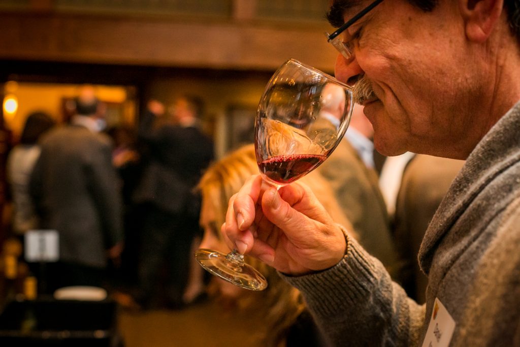A man holds a wineglass up to his nose, with a blurry crowd in the background.