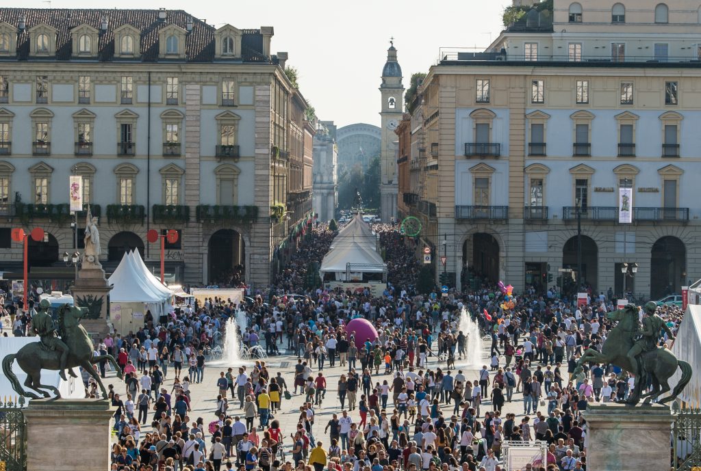 A colorful crowd moves through a city plaza with temporary white tents and water fountains.