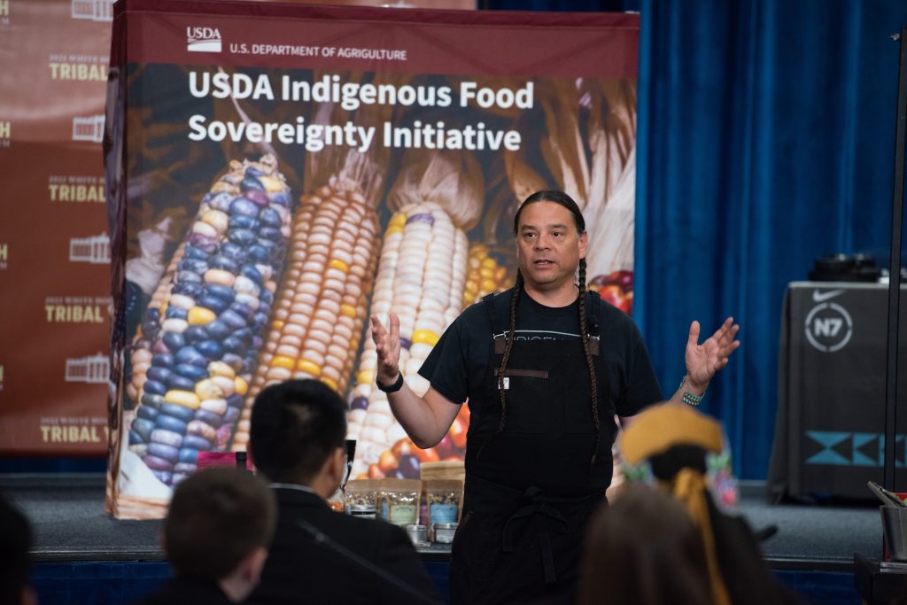 A man with two long braids wearing a chef’s apron speaks to an audience, in front of a banner for the USDA Indigenous Food Sovereignty Initiative.