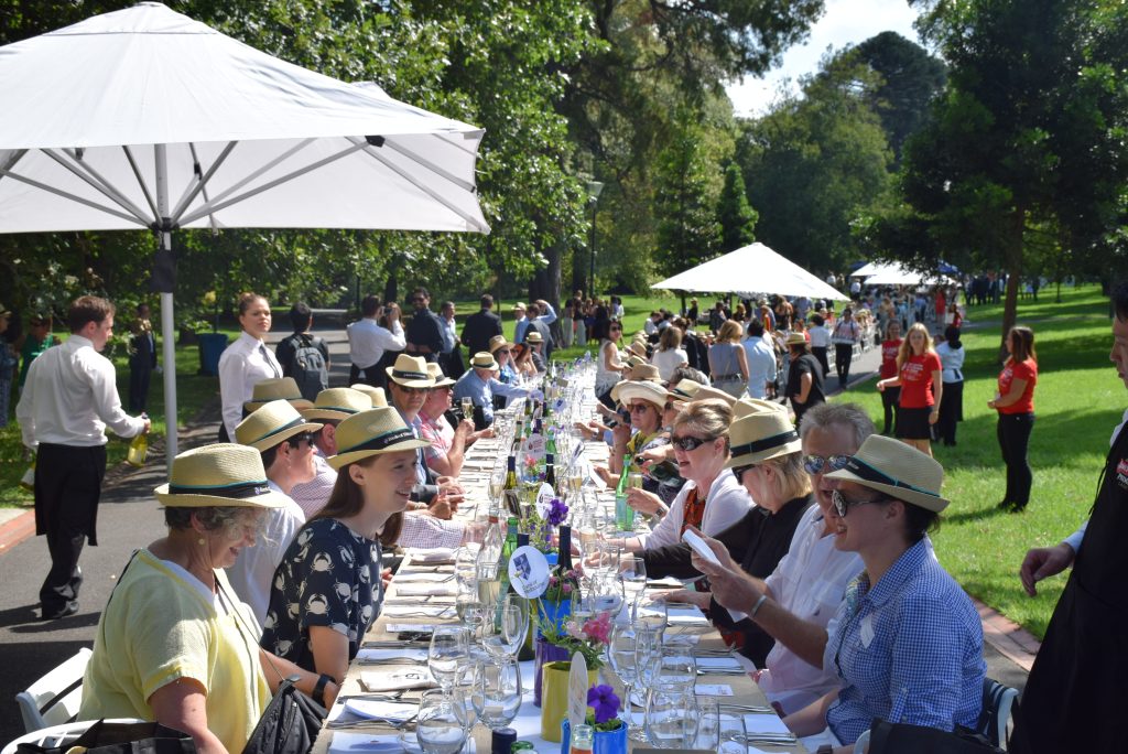 A crowd dressed for a hot day, many wearing straw fedoras, sit on either side of a long lunch table that extends into the distance on a walkway in a park.