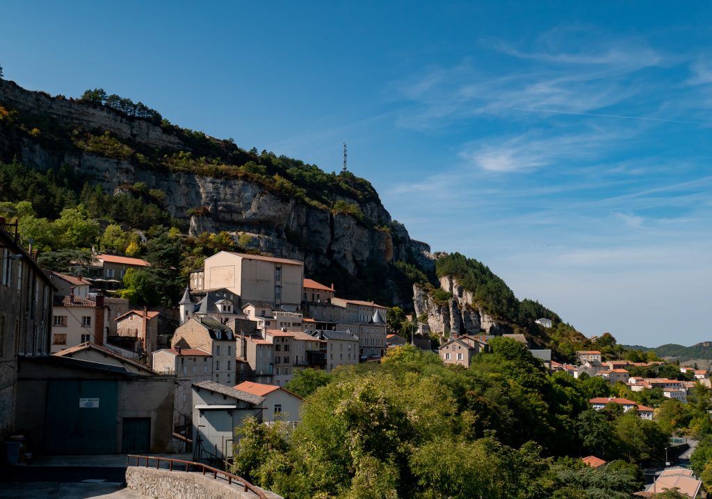 Tan and white buildings, interspersed with a variety of green trees, spread out at the foot of a short cliff.
