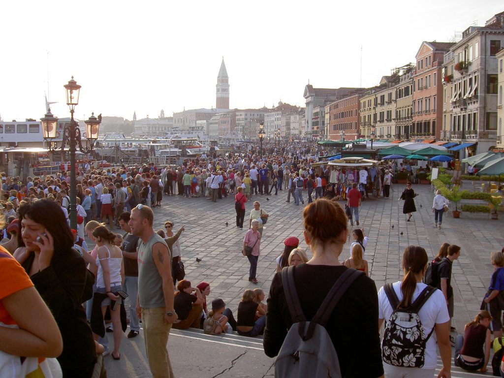 A street-level photo of a crowded cobblestone street, flanked by buildings and canvas awnings on one side, and sightseeing boasts on the other.