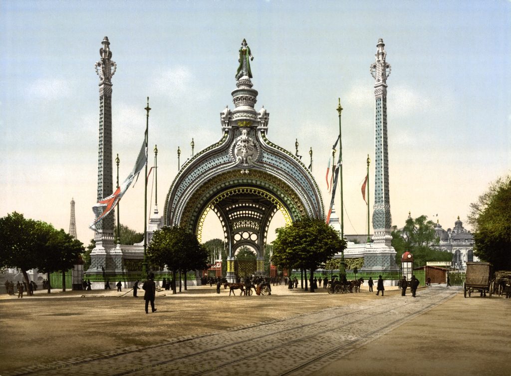A towering white arch with almost lace-like details is on the far side of a plaza with a few horse-drawn carriages and walking figures in black suits. The Eiffel Tower is visible in the far distance.