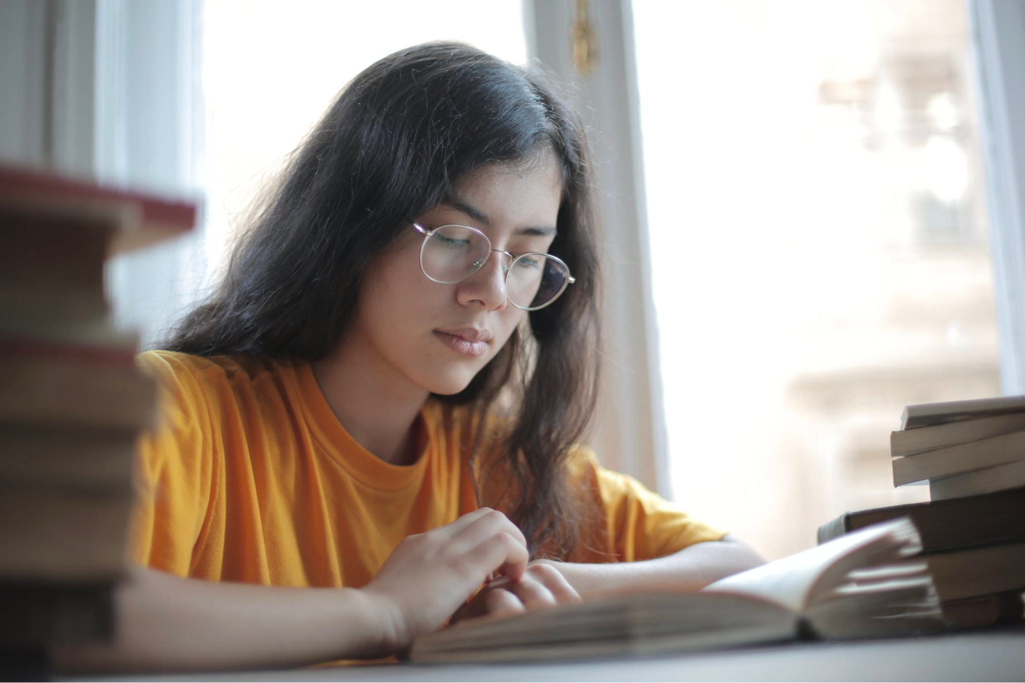 A young woman is at a table reading a book with more books piled around her