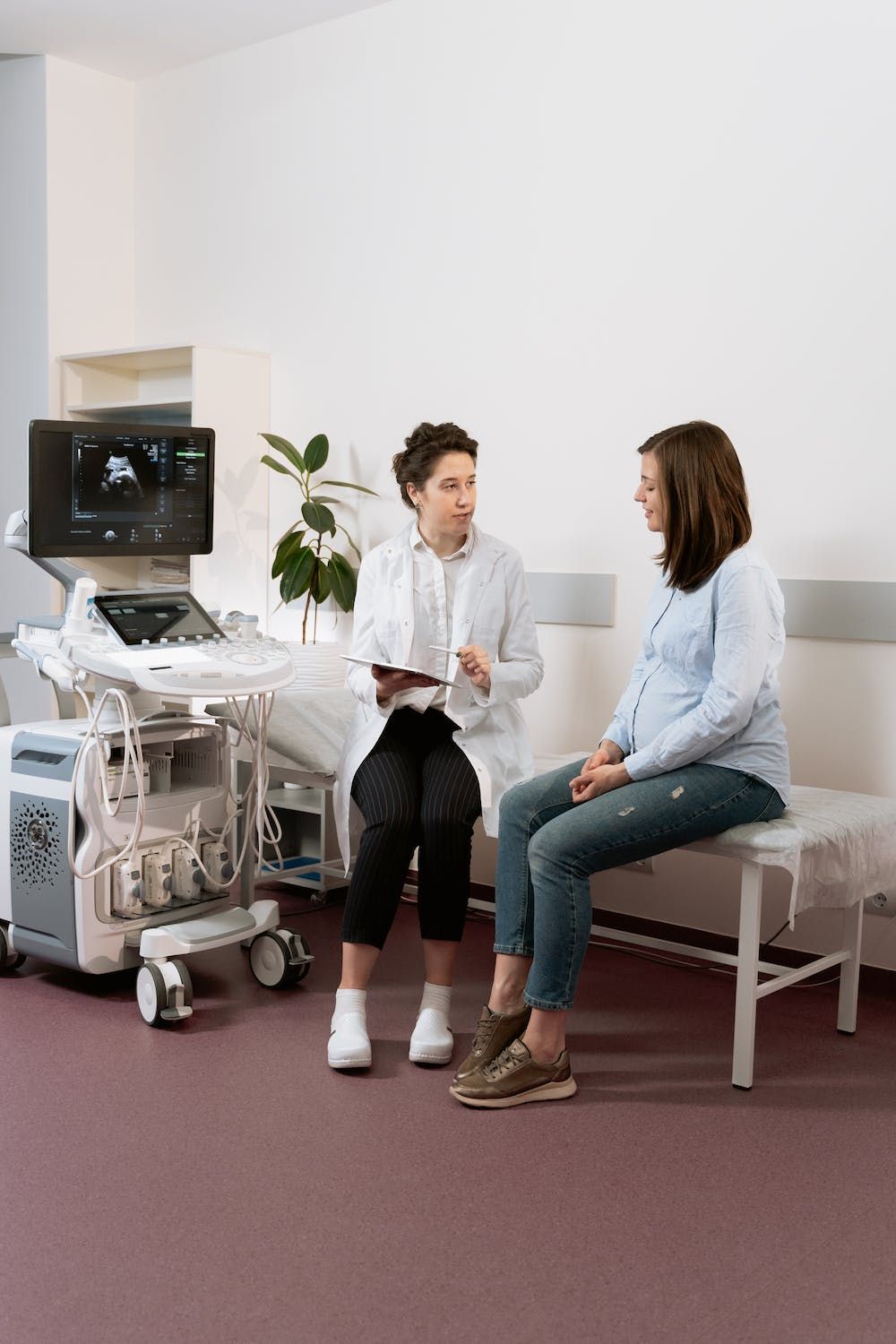 A health professional in a white coat sits next to a pregnant woman on an exam table. There is an ultrasound machine in the room.